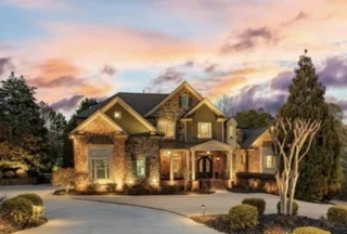 A large two-story brick and stone house is lit at dusk under a colorful sunset, with a curved driveway and manicured landscaping.